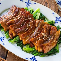 Honey pork belly slices topped with sesame seeds, served over saut&eacute;ed greens on a patterned oval platter.
