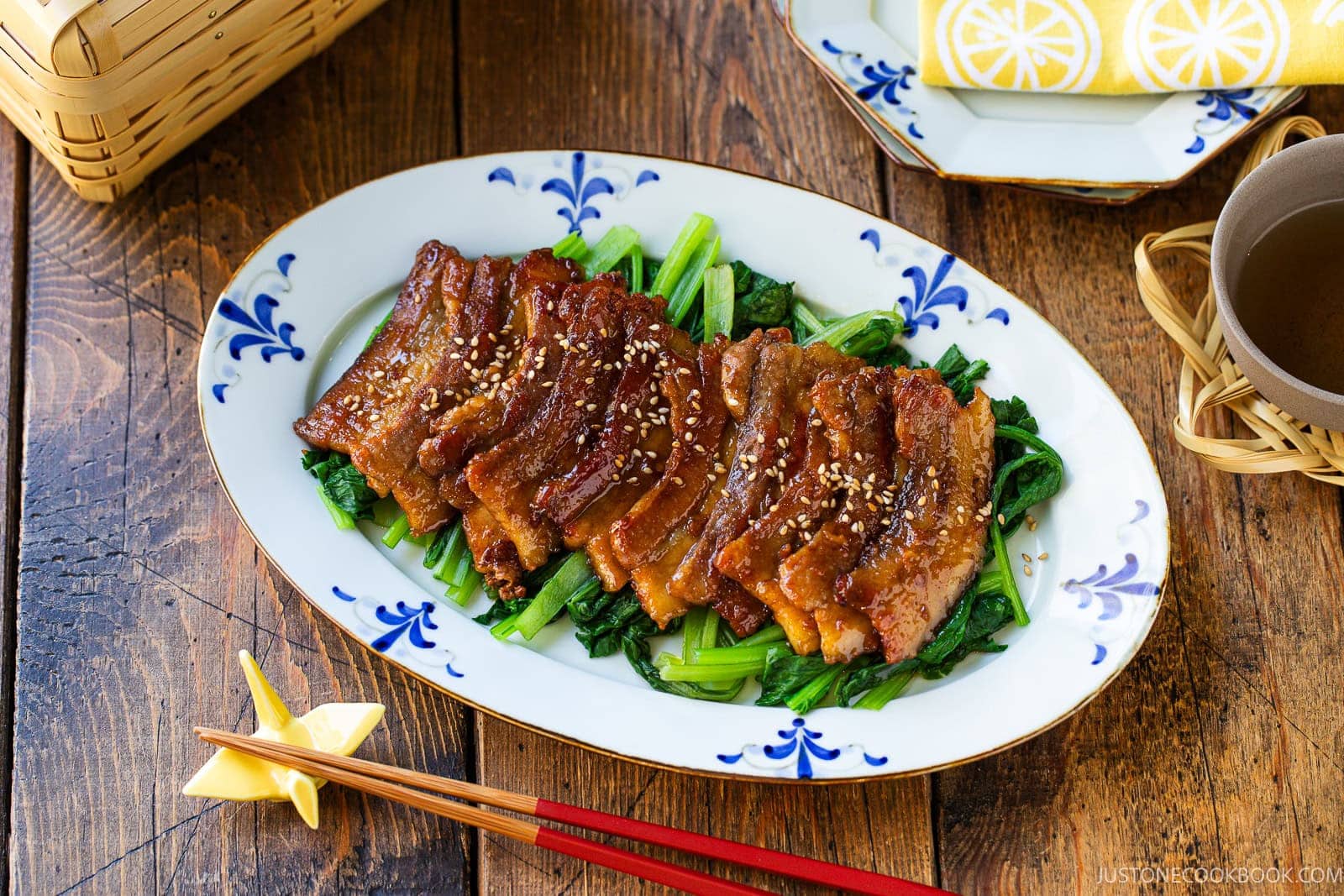 Honey pork belly slices topped with sesame seeds, served over saut&eacute;ed greens on a decorative oval platter with chopsticks nearby.