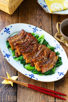Honey pork belly slices glazed and topped with sesame seeds, served over saut&eacute;ed greens on a patterned oval plate.