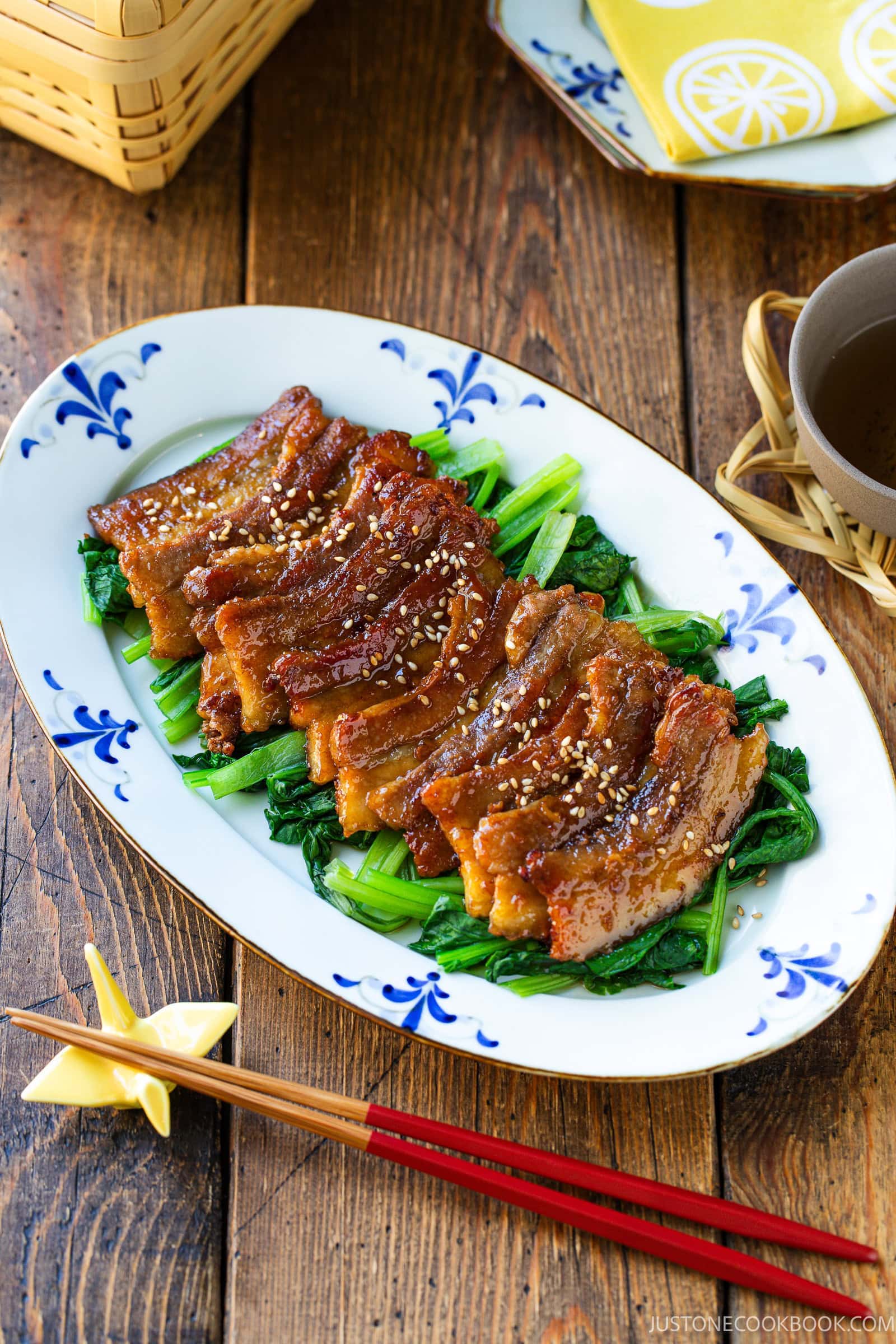 Honey pork belly slices glazed and topped with sesame seeds, served over saut&eacute;ed greens on a patterned oval plate.