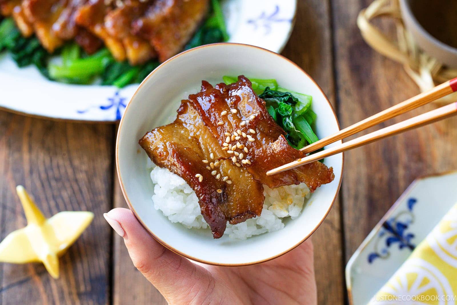 A hand holds a small bowl of white rice topped with honey pork belly and greens, with chopsticks lifting a piece of the meat.