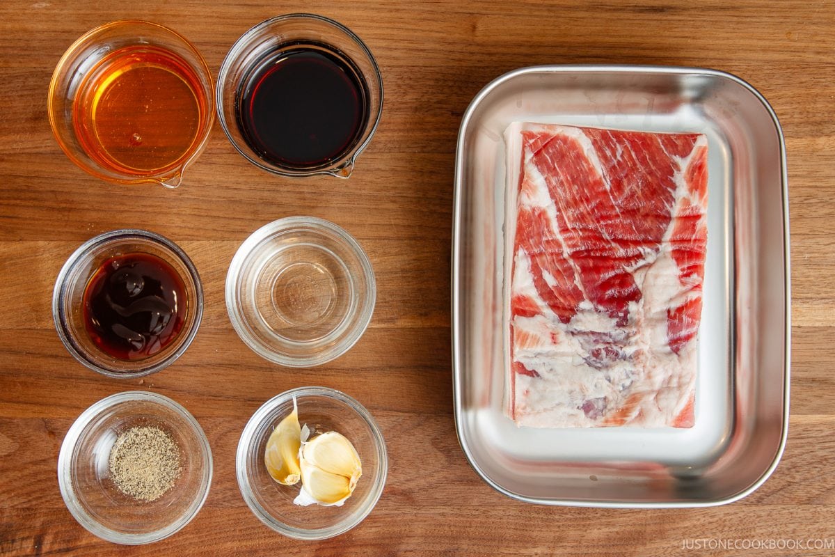 A rectangular slab of raw honey pork belly in a metal tray alongside small bowls containing soy sauce, oyster sauce, sake, ground pepper, and two garlic cloves.