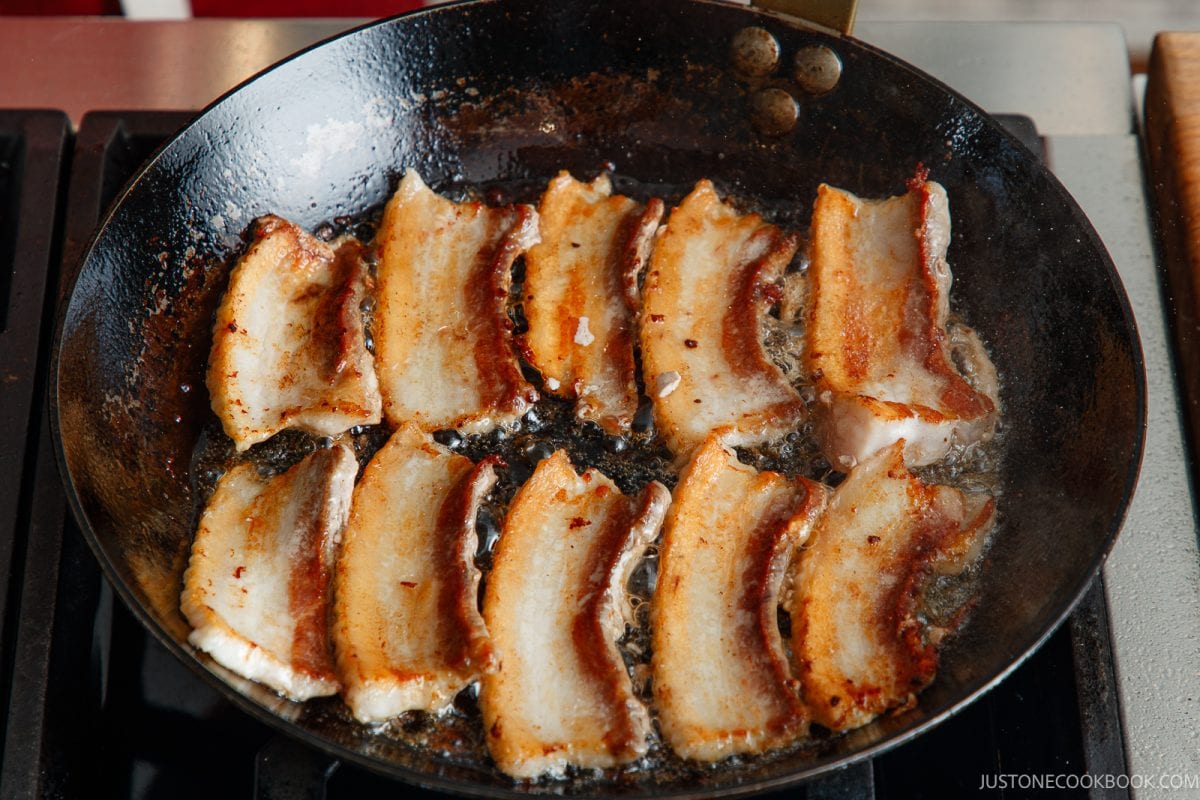 Slices of honey pork belly frying in a carbon steel skillet.