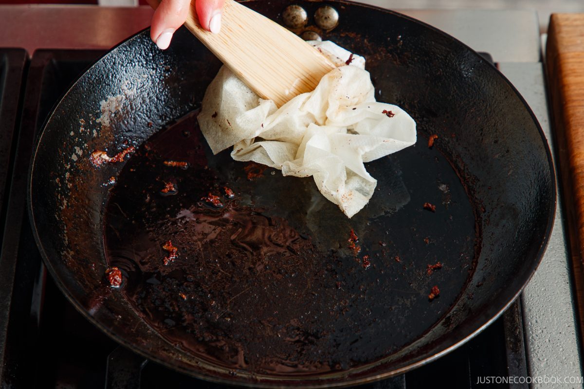 A hand uses a wooden spatula to wipe the inside of a dark, oily skillet used for honey pork belly with a folded paper towel.
