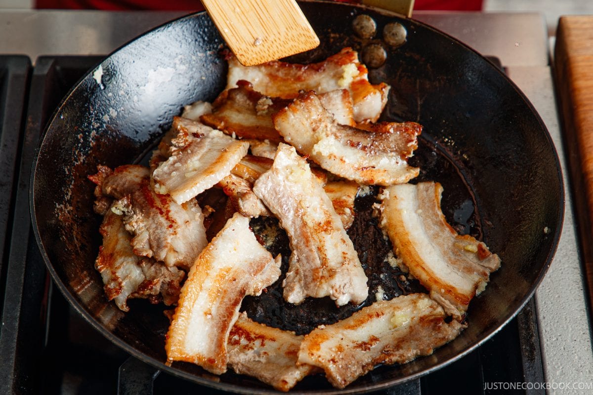 Slices of honey pork belly frying in a skillet being moved by a wooden spatula.