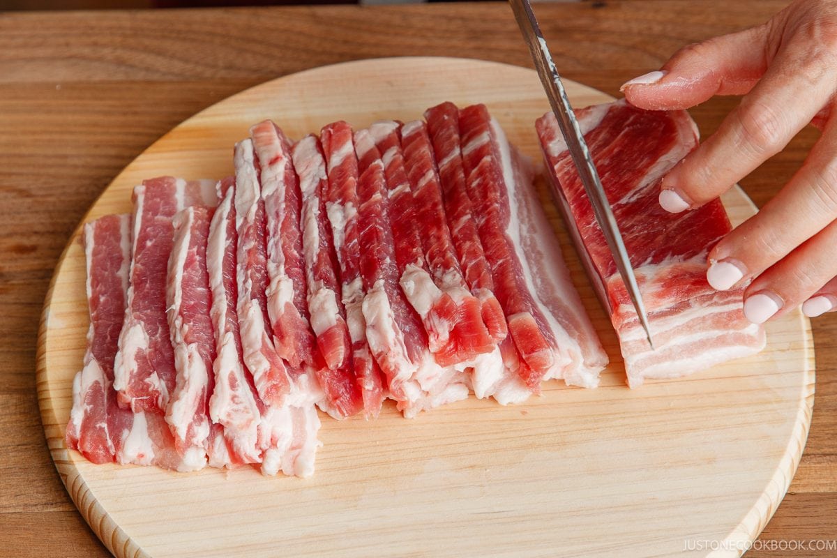 A knife slices a slab of honey pork belly on a wooden cutting board next to several thin, pre-cut strips of meat.