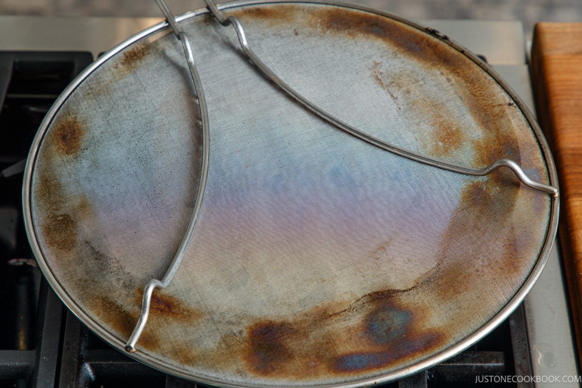 Round metal grease splatter screen sitting on a stovetop while cooking honey pork belly.