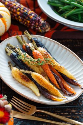 A plate of miso glazed roasted carrots and multicolored baby carrots garnished with herbs, placed on a rustic wooden table set with gold utensils, green beans, Indian corn, and fall-themed decor in the background.