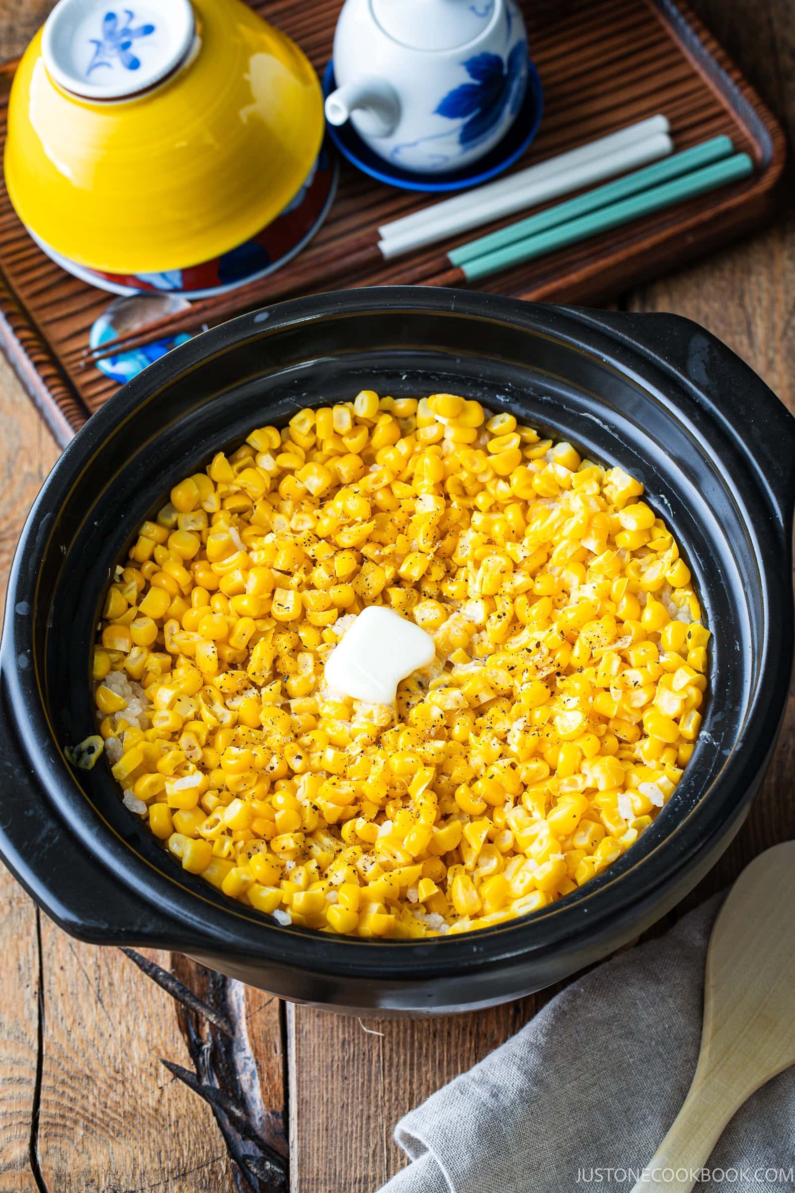 A black pot filled with yellow corn kernels, a pat of butter, and black pepper sits on a wooden table next to bowls, cups, and chopsticks—an inviting presentation reminiscent of Japanese corn rice.