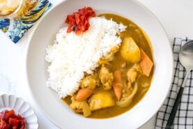 A plate of Japanese chicken curry with white rice, topped with red pickles. The curry contains chunks of chicken, carrots, and potatoes. A spoon and napkin are placed to the right of the plate.