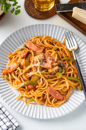 A plate of ketchup spaghetti with sausage, green peppers, onions, and mushrooms, topped with grated cheese, sits on a white dish with a fork on the side.