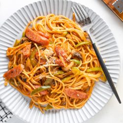 A plate of ketchup spaghetti with tomato sauce, sliced sausage, green bell peppers, and mushrooms, topped with grated cheese. A fork rests on the plate next to a drink, napkin, and cheese grater.