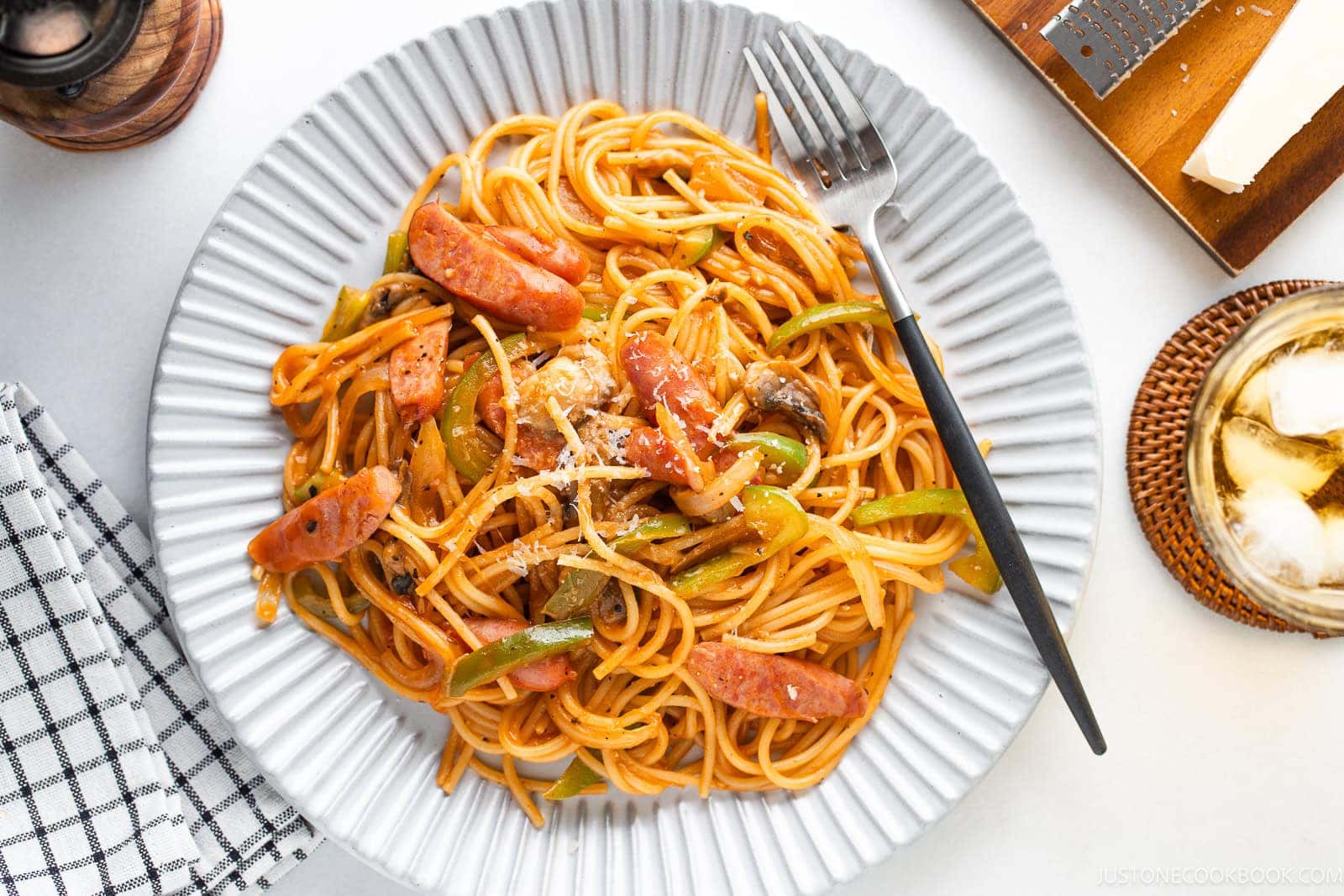 A plate of Napolitan (ketchup spaghetti with tomato sauce), sliced sausage, green bell peppers, and mushrooms, topped with grated cheese. A fork rests on the plate next to a drink, napkin, and cheese grater.
