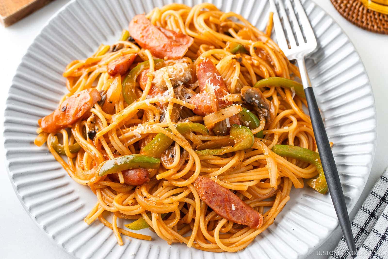 A Napolitan (ketchup spaghetti with tomato sauce) with sliced sausages, bell peppers, mushrooms, and tomato sauce is served with a fork on a light-colored, ridged plate.