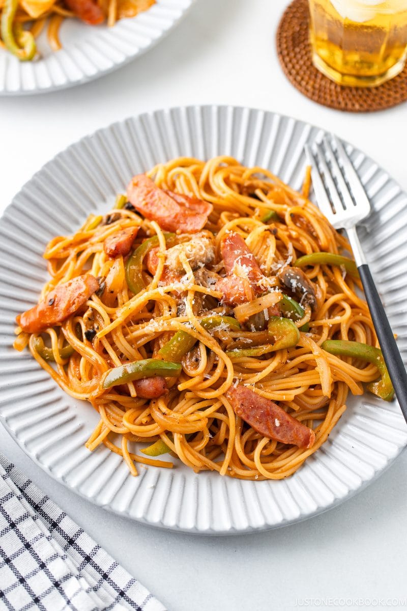 A plate of Napolitan (ketchup spaghetti with tomato sauce) with sliced sausage, green bell peppers, onions, and a tangy tomato-based sauce, served with a fork. A glass of iced drink and another plate are visible in the background.