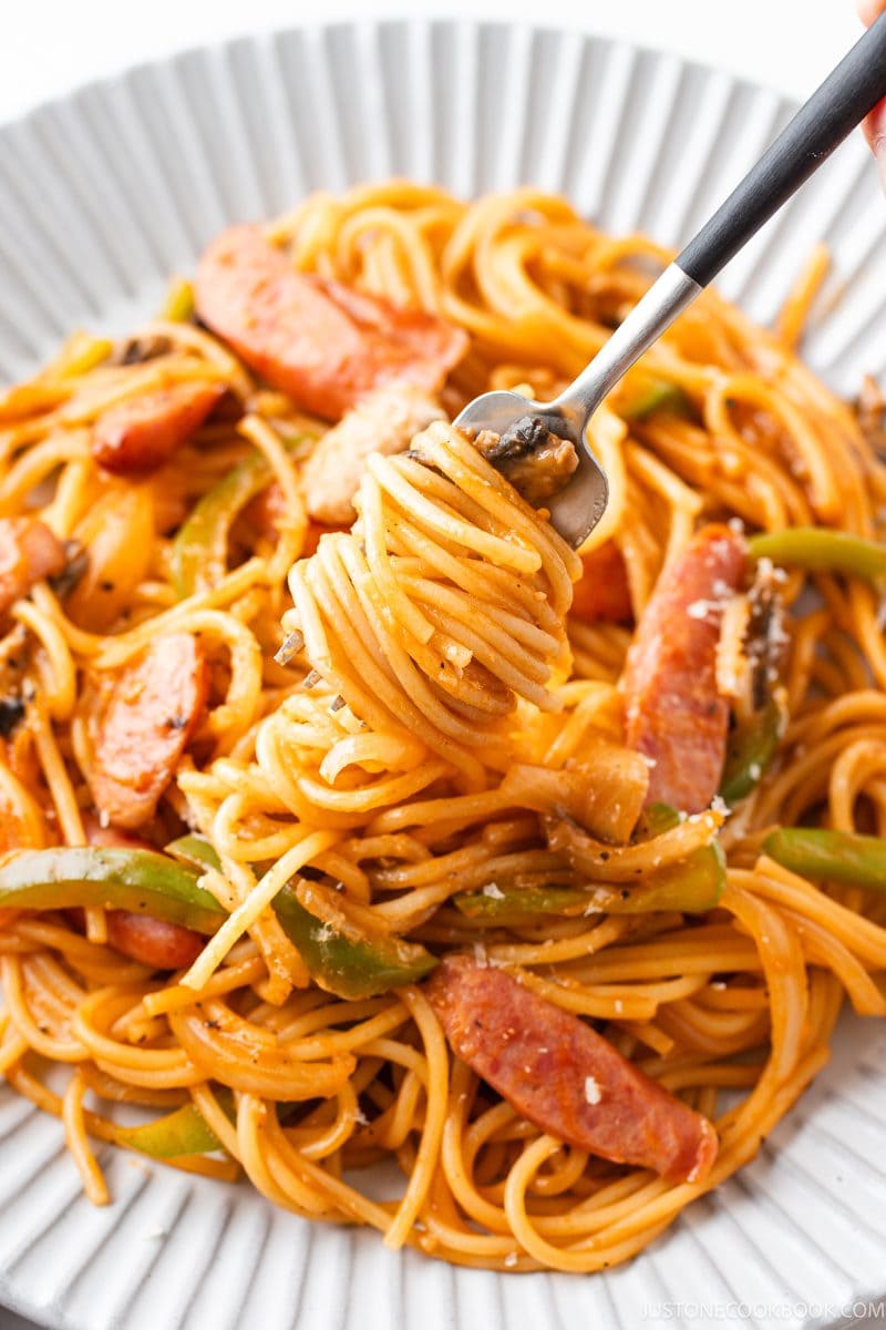 A plate of ketchup spaghetti with sausage, green peppers, onions, and tomato sauce, with a fork lifting a twirl of pasta.