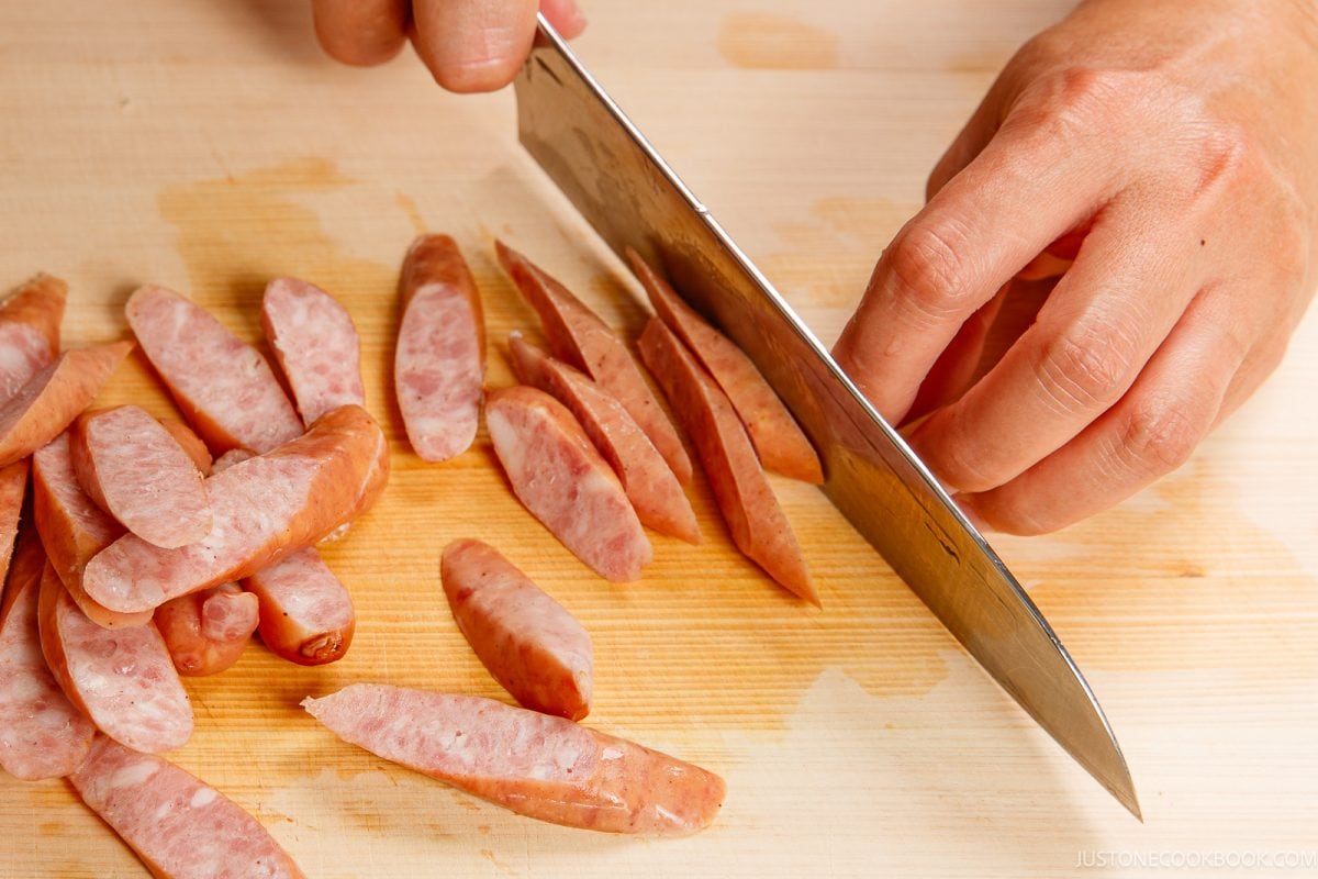 A person slices sausage links into thin pieces on a wooden cutting board with a large kitchen knife, preparing the ingredients for ketchup spaghetti.
