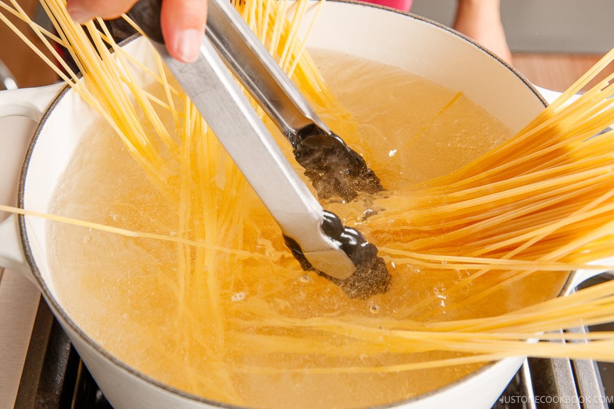 A hand uses tongs to stir uncooked ketchup spaghetti in a large pot of boiling water on a stove.