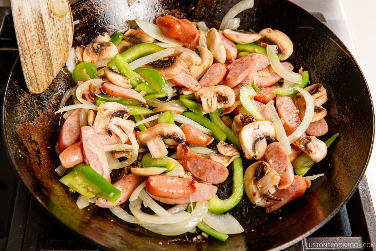 Sliced sausage, mushrooms, green bell peppers, and onions are being stir-fried in a large pan on a stovetop to create a delicious base for ketchup spaghetti. A wooden spatula rests on the side of the pan.