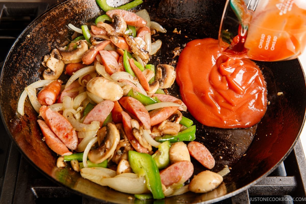 Sliced sausages, onions, mushrooms, and green peppers cooking in a skillet evoke the classic flavors of ketchup spaghetti, while tomato sauce is being poured in from a measuring cup on the right side.