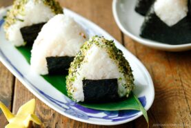Three classic onigiri rice balls wrapped with seaweed and sprinkled with seasoning sit on a plate with a green leaf, set on a wooden table. More delicious onigiri are arranged on a plate in the background.