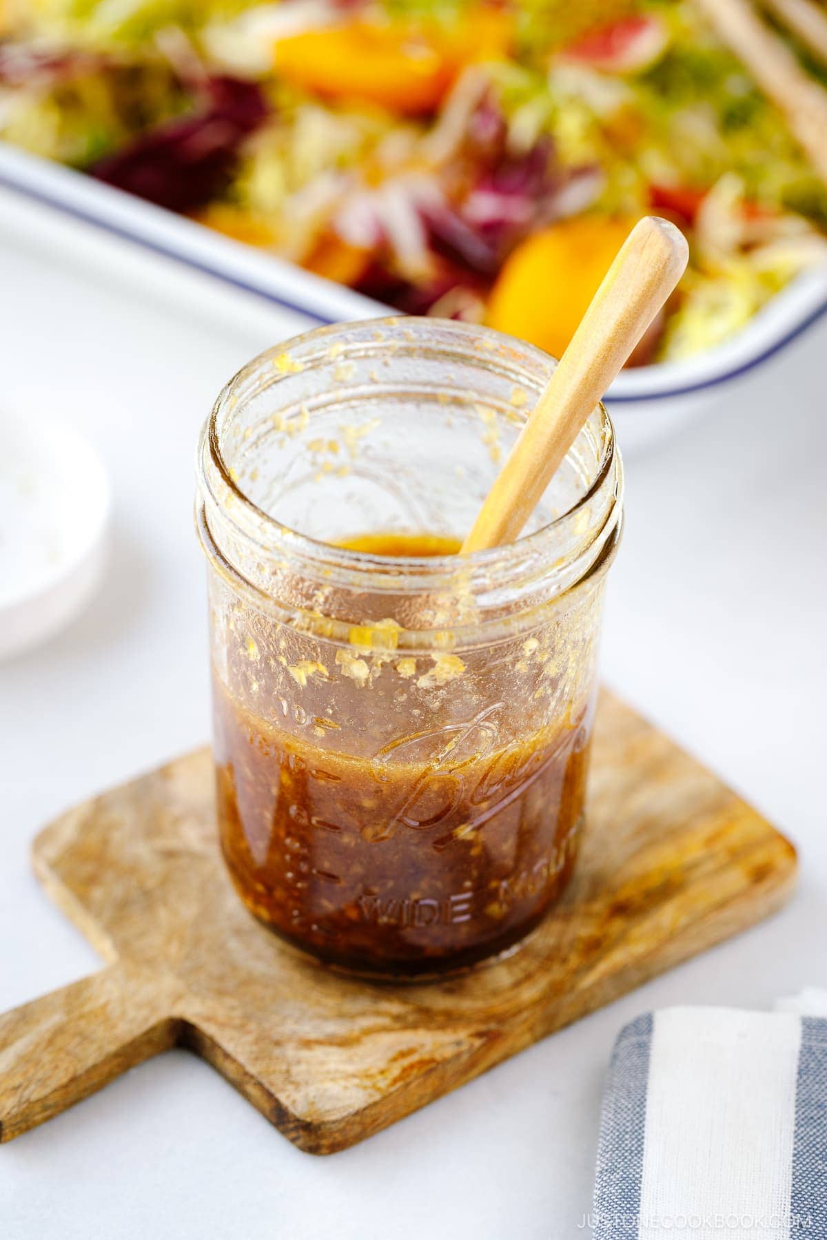 A glass jar filled with brown Japanese onion dressing sits on a small wooden board, with a wooden spoon inside. A colorful salad is blurred in the background.