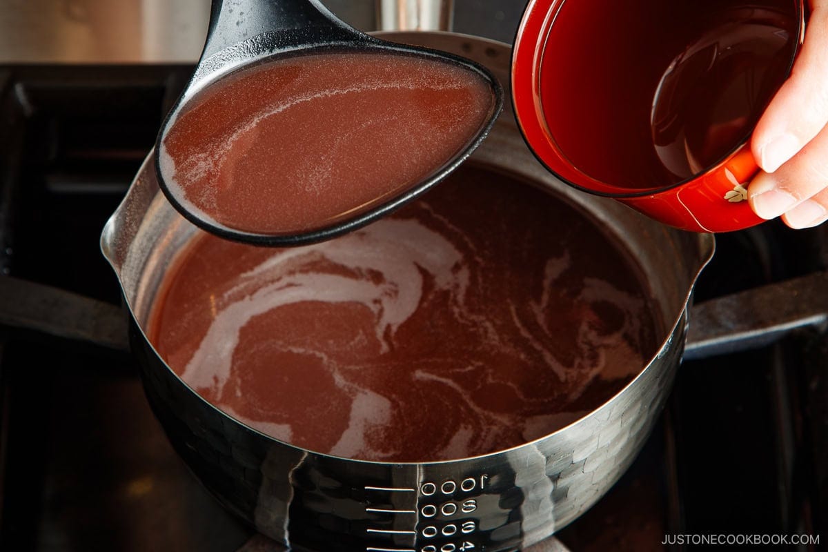 A hand pours liquid from a red bowl into a saucepan of oshiruko, or sweet red bean soup, while a ladle scoops some soup, revealing the smooth, maroon surface with a swirl.