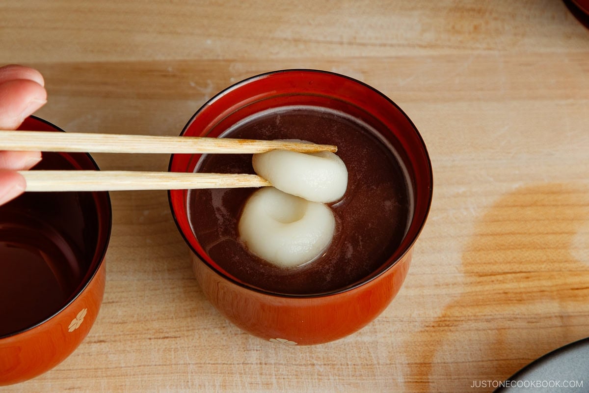 A hand using chopsticks to pick up a round, white mochi from a bowl of sweet red bean soup, known as oshiruko, on a wooden surface.