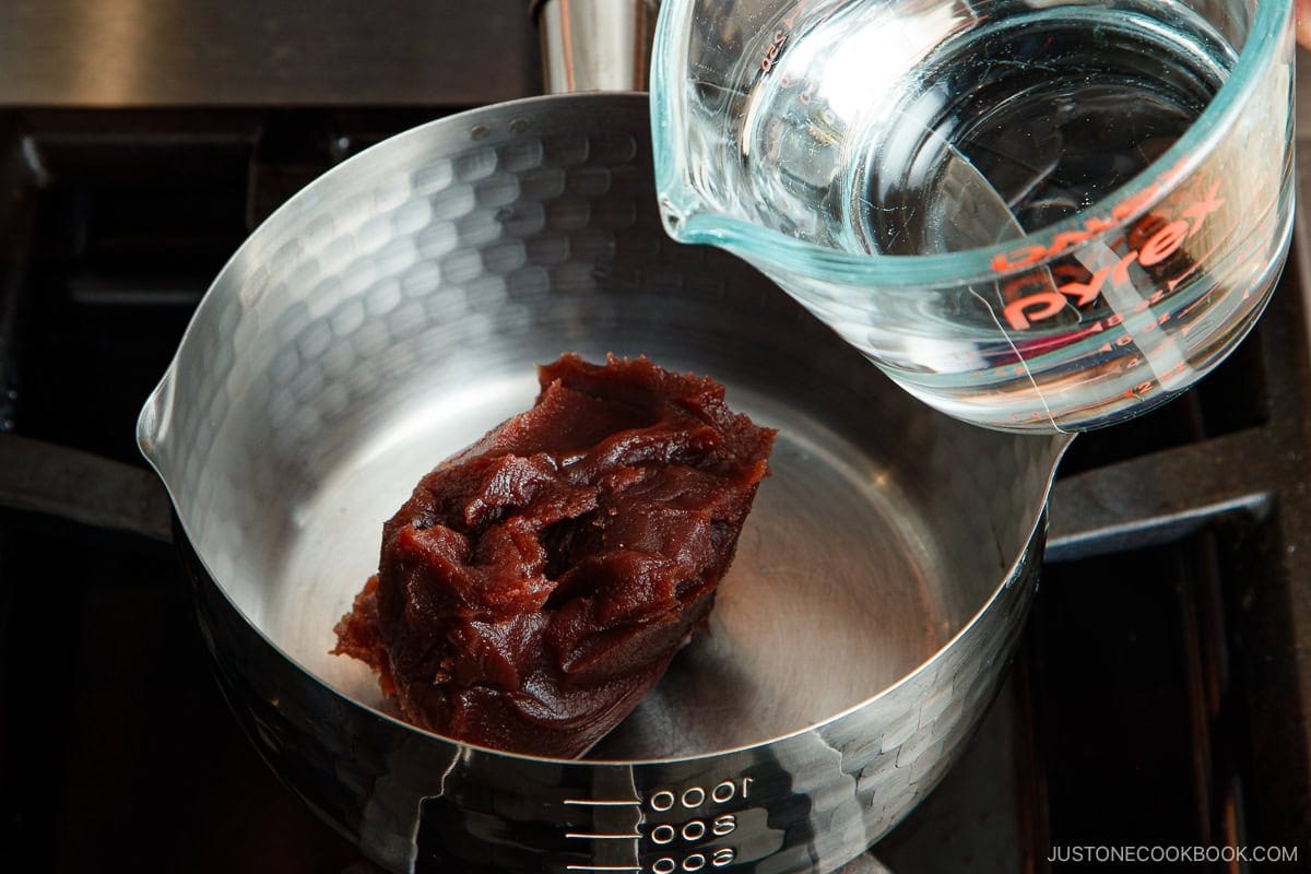 A pot with a scoop of red bean paste inside, placed on a stove, while a measuring cup of water is being poured in&mdash;perfect for preparing comforting dishes like oshiruko.