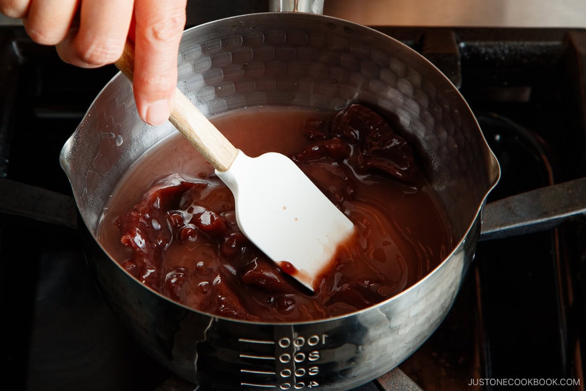 A hand stirs red bean paste with a white spatula in a silver pot on a stovetop, preparing oshiruko.