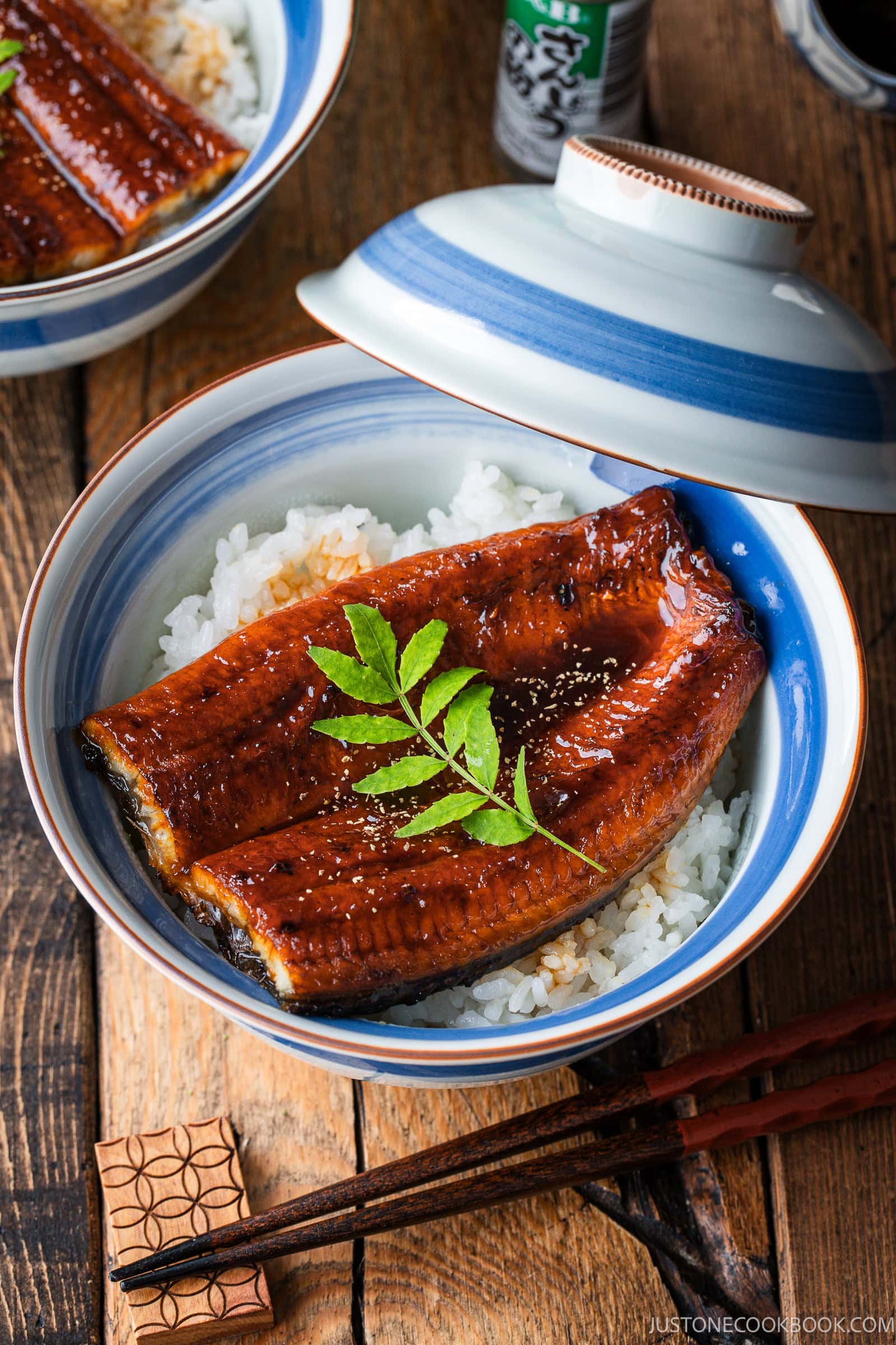 Unadon of glazed grilled eel served over white rice in a ceramic bowl with a lid, garnished with a sprig of sansho pepper leaves.