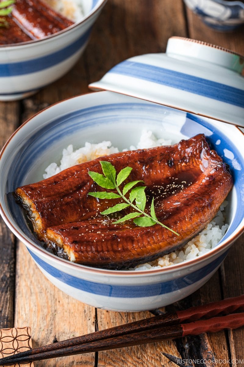 A bowl of unadon over white rice, garnished with a green leaf and served with a matching lid on a wooden table.