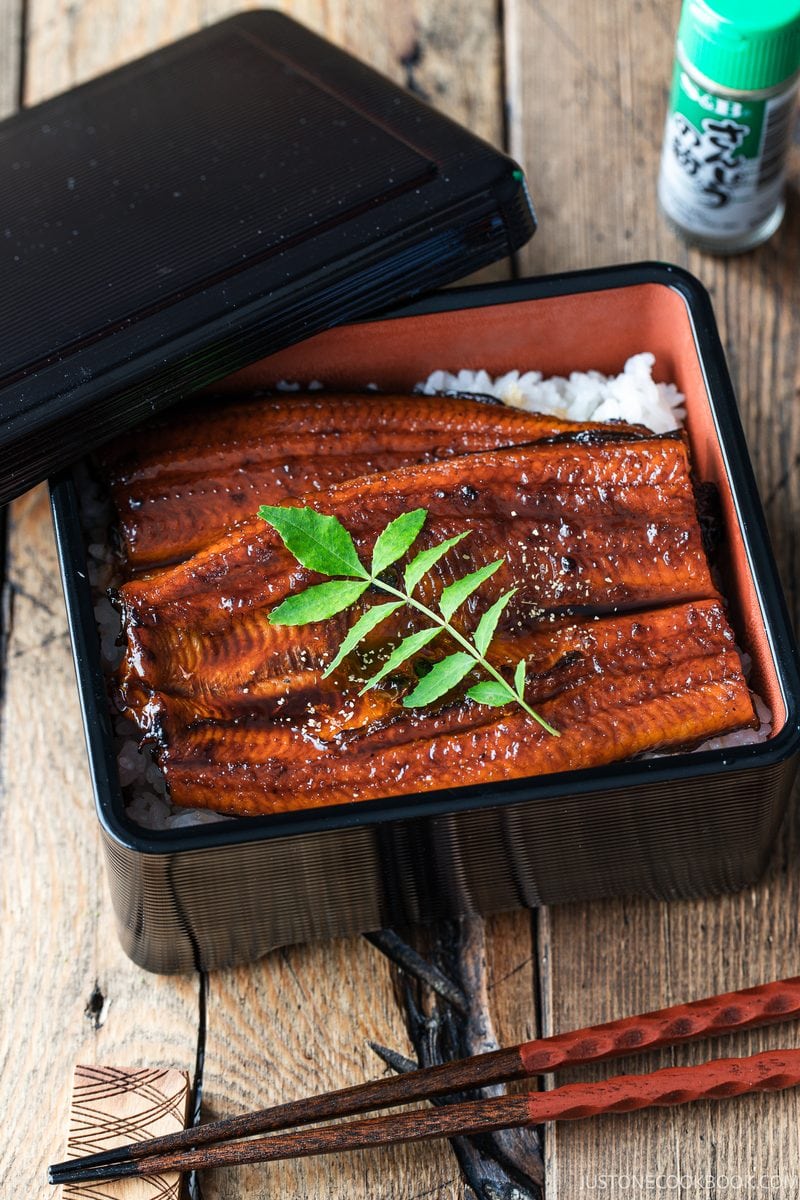 A black lacquered box containing unadon, featuring grilled eel over white rice topped with a green leaf, with chopsticks in the foreground.