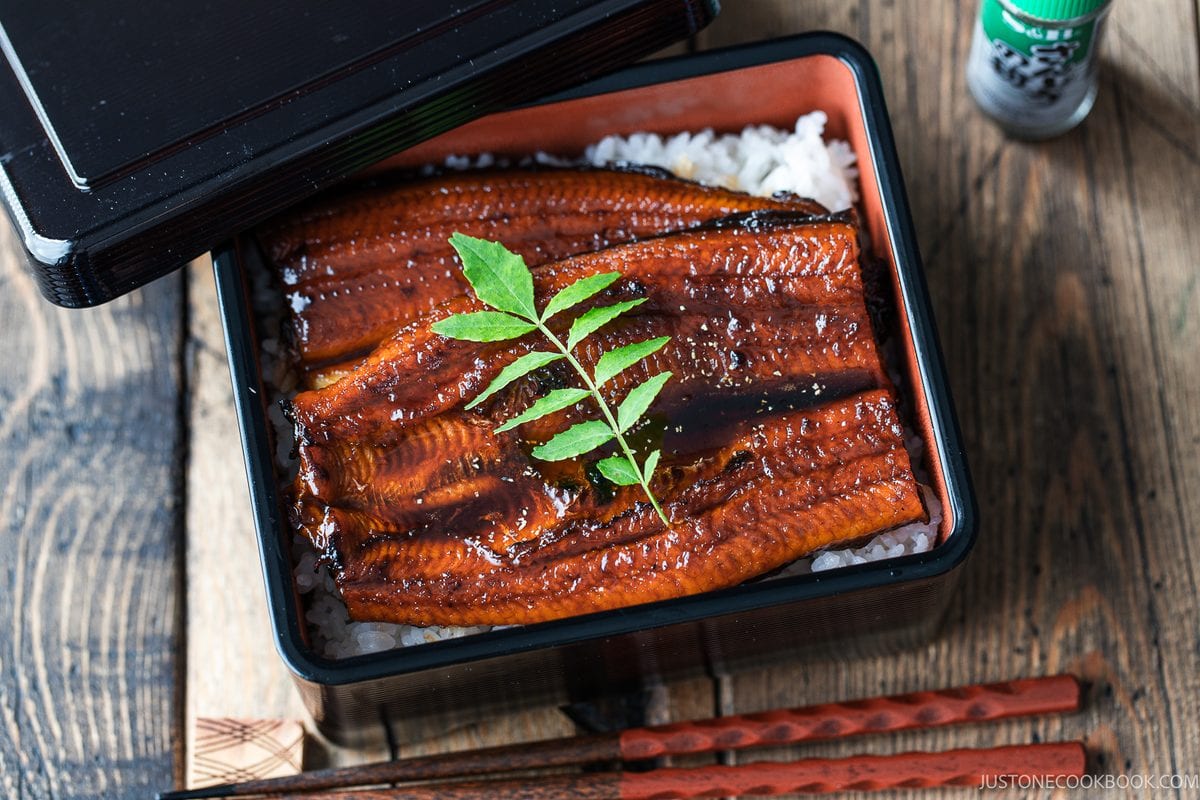 A black bento box containing unadon, grilled unagi eel glazed in sauce over white rice, garnished with a sprig of sansho leaf.