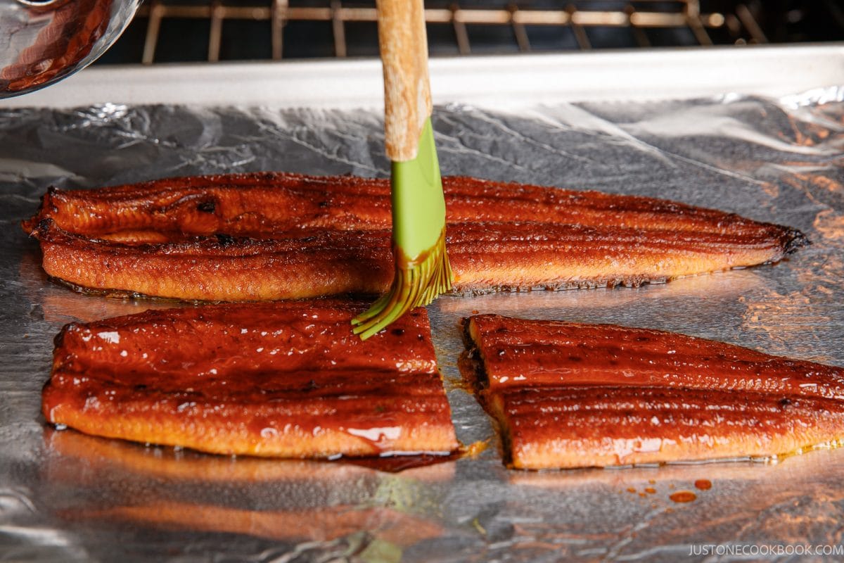 A silicone brush glazes unadon roasted eel fillets on a foil-lined baking sheet.