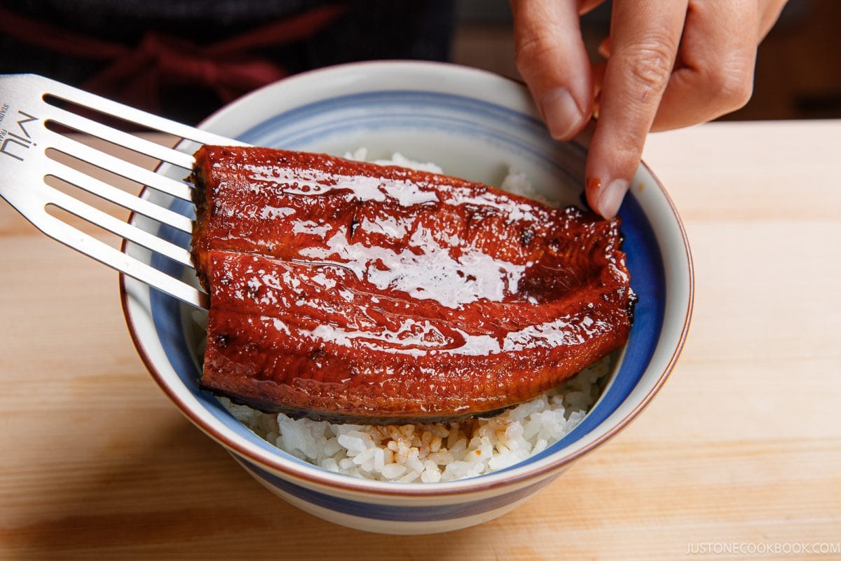 A person uses a metal spatula to place a glossy, glazed piece of grilled eel over a bowl of white rice to serve unadon.