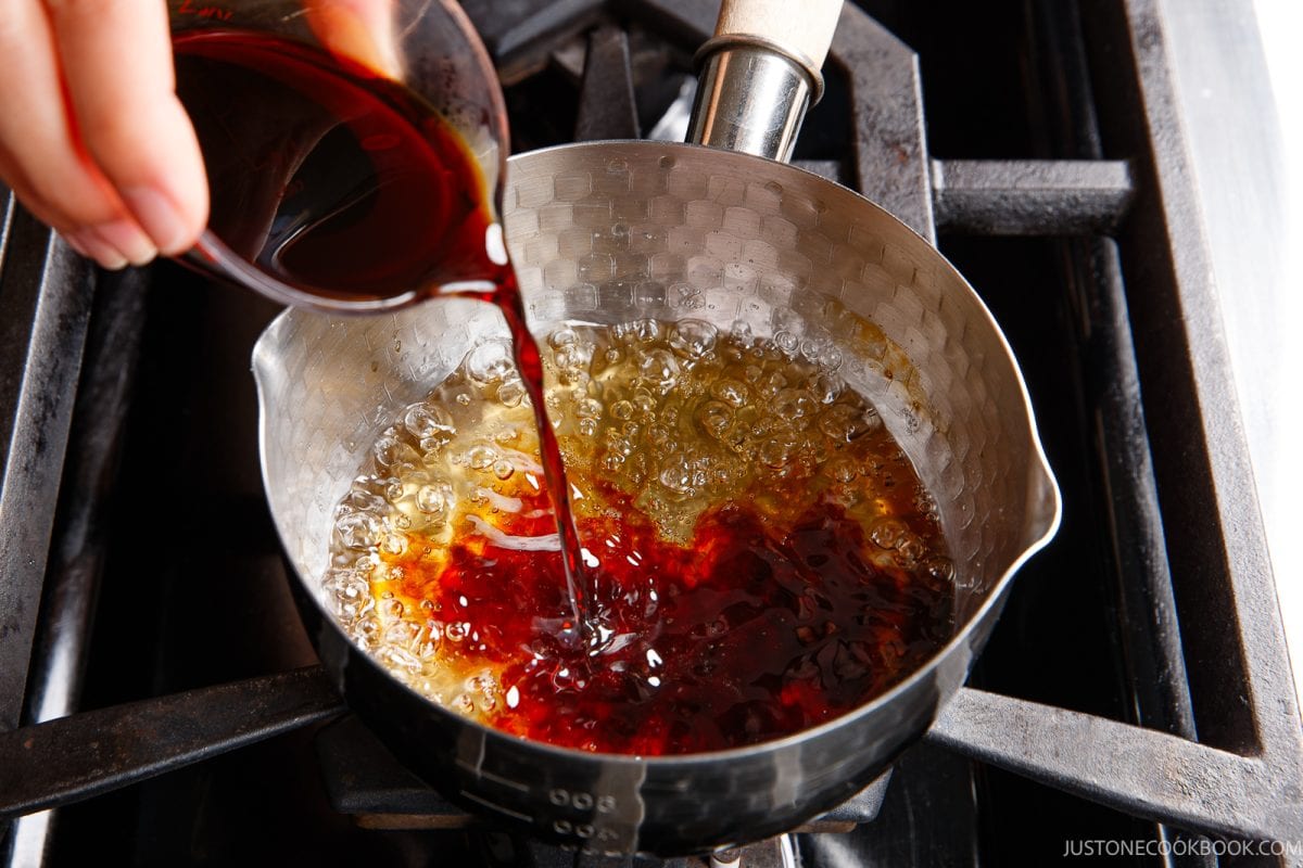 Dark unadon sauce is poured from a measuring cup into a small saucepan of bubbling golden liquid on a stove.