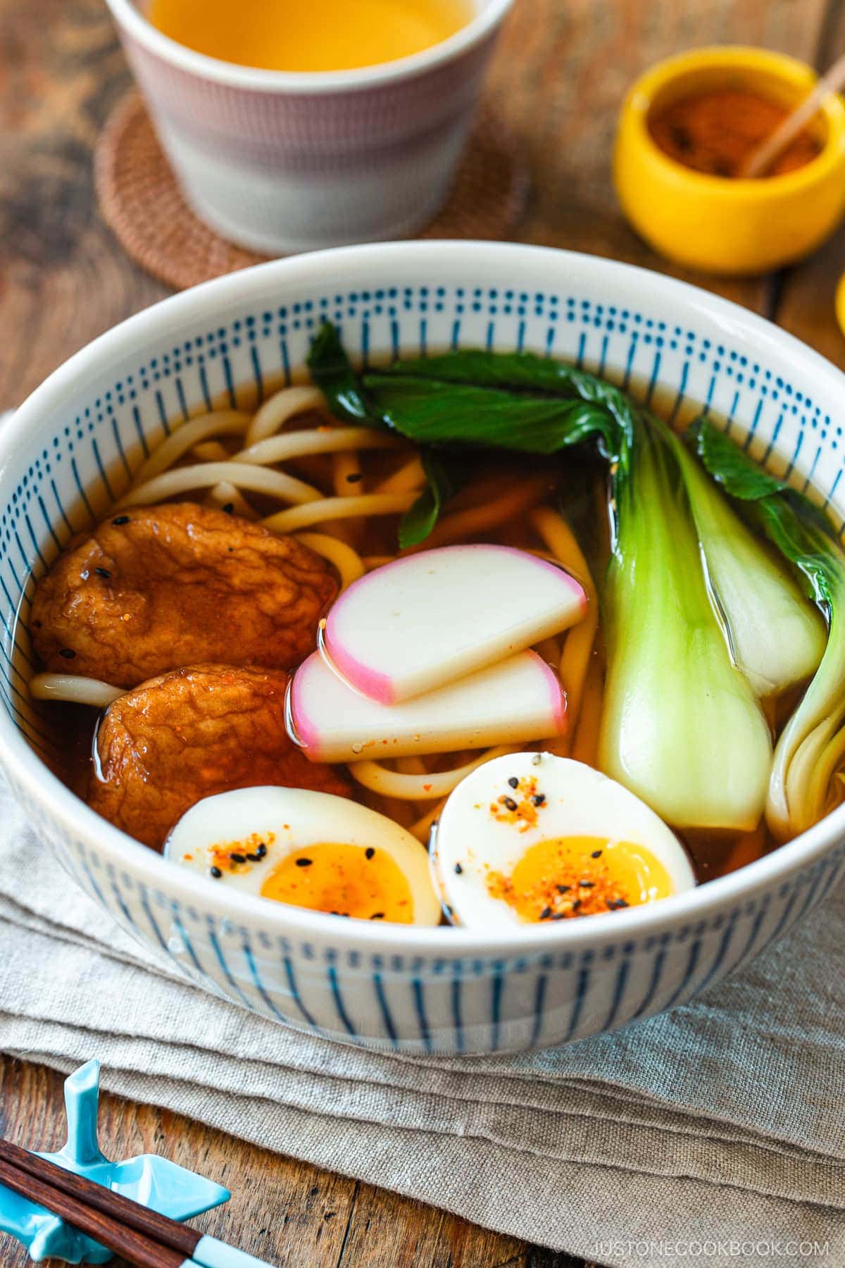 A large blue and white bowl containing udon noodle soup topped with fish cake, bok choy, fish cakes, and jammy soft boiled eggs in a savory dashi broth.