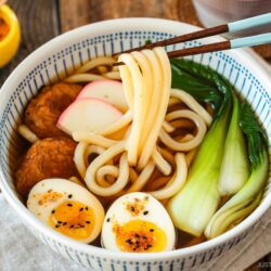 A large blue and white bowl containing udon noodle soup topped with fish cake, bok choy, fish cakes, and jammy soft boiled eggs in a savory dashi broth.