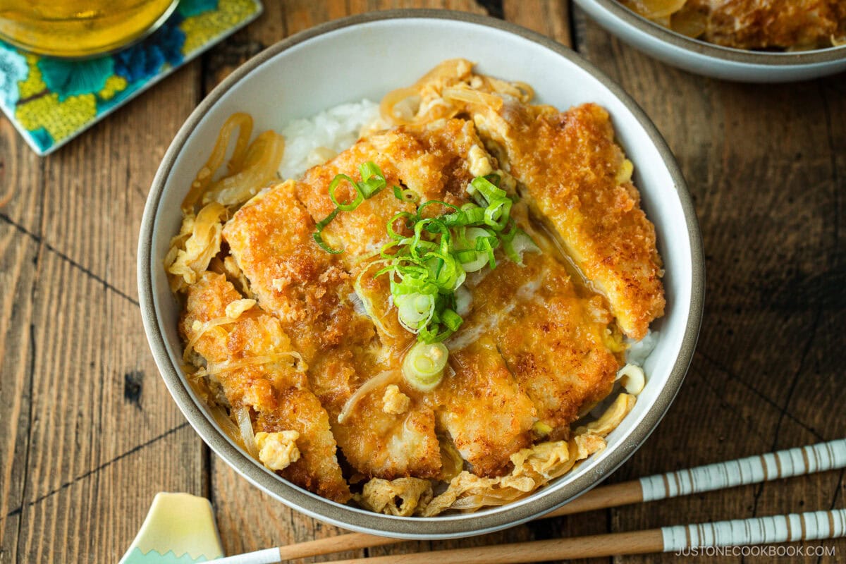 A white bowl containing Katsudon, pork cutlet rice bowl, where golden brown tonkatsu, drizzled egg, and tender onion are simmered in a savory dashi broth.