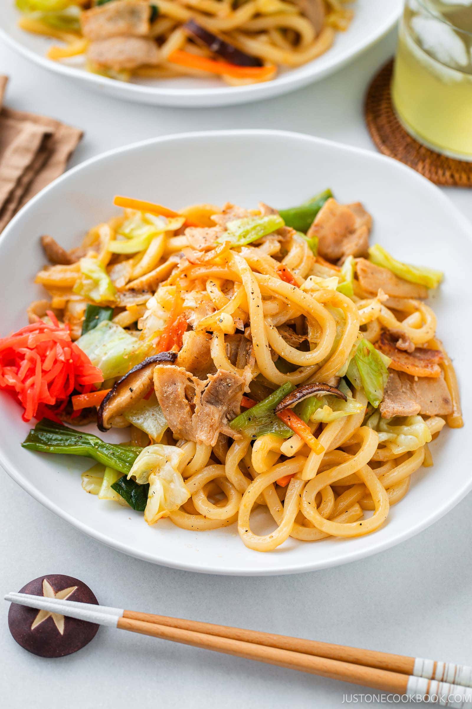 A white plate of savory yaki udon noodles with pork, vegetables, pickled ginger, and sesame seeds. A pair of chopsticks and a glass of iced green tea are beside the plate.