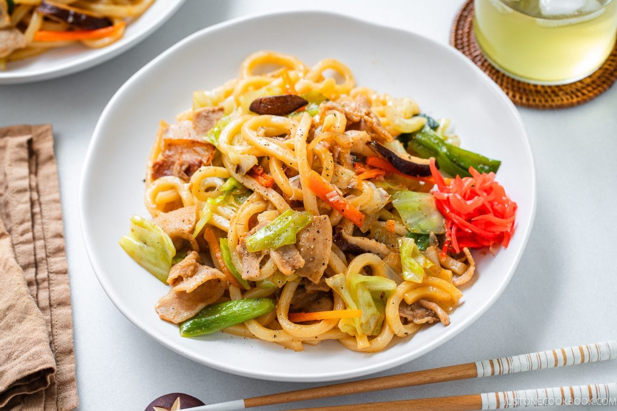 A plate of yaki udon stir-fried noodles with sliced chicken, cabbage, carrots, green onions, and pickled ginger is served with a glass of iced drink and chopsticks on a white table.