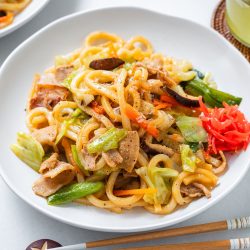 A plate of yaki udon stir-fried noodles with sliced chicken, cabbage, carrots, green onions, and pickled ginger is served with a glass of iced drink and chopsticks on a white table.