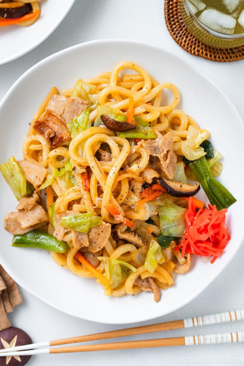 A steaming plate of yaki udon features thick noodles stir-fried with sliced pork, cabbage, carrots, mushrooms, and tangy pickled ginger, served with chopsticks and a refreshing glass of iced tea nearby.
