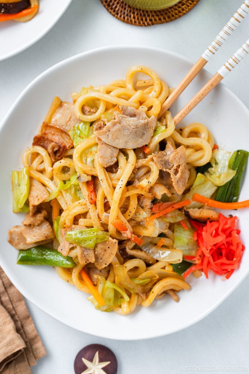 A delicious bowl of yaki udon featuring thick noodles, savory pork slices, fresh vegetables, and pickled ginger, beautifully garnished and served with chopsticks.