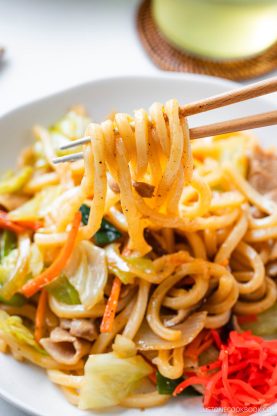 Chopsticks holding a bite of stir-fried yaki udon noodles with vegetables, including cabbage and carrots, over a plate of savory yaki udon. Pink pickled ginger is on the side.