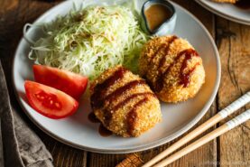 A round white plate containing Japanese croquettes, shredded cabbage, tomato wedges, and a tiny container with sesame dressing.