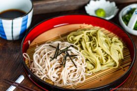 A red tray of Zaru Soba (Cold Soba) features white and green soba noodles topped with sliced seaweed, placed on a bamboo mat beside a bowl of dipping sauce, wasabi, and chopped scallions. Chopsticks rest alongside the tray.