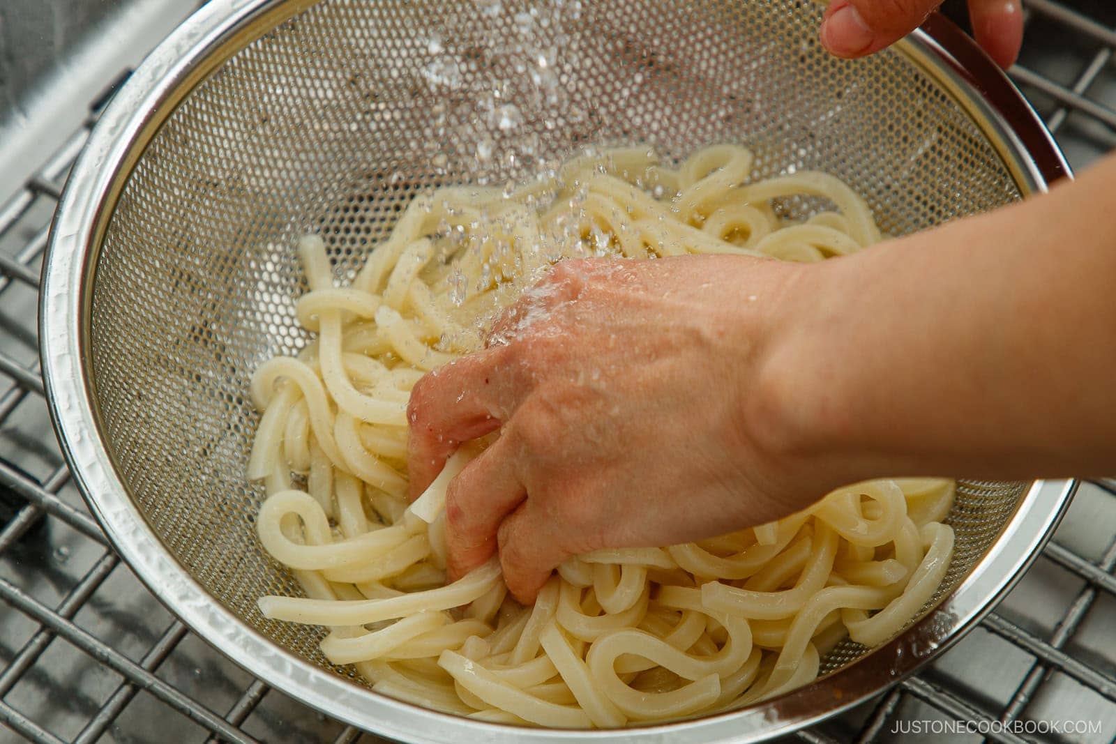 A hand rinses and scrubs cooked udon noodles in a metal strainer under running water, preparing Zaru Udon (Cold Udon) in a kitchen sink.