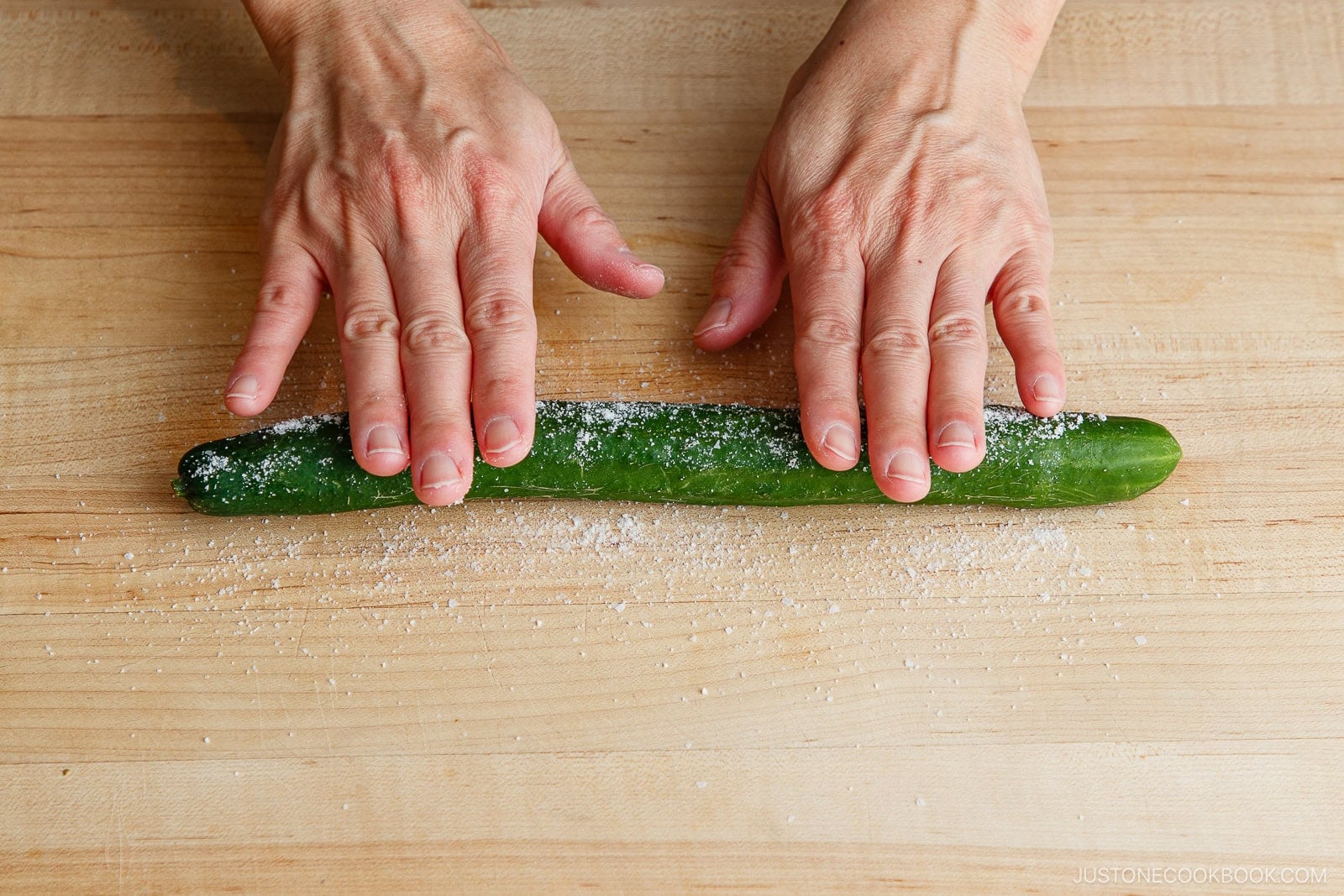 Two hands doing prep work, rolling a cucumber on a wooden surface sprinkled with salt.